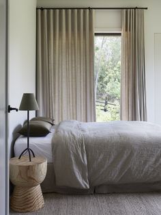 a bed sitting under a window next to a wooden table with a lamp on it