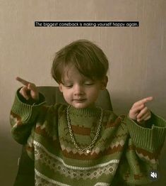 a young boy sitting in a chair wearing a green sweater and pointing to the side