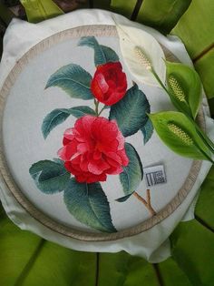 a red flower painted on a white cloth with green leaves surrounding it and a small round wooden frame in the center