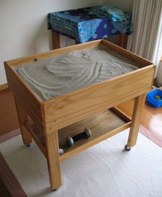 a wooden table with sand in it on top of a carpeted floor next to a window