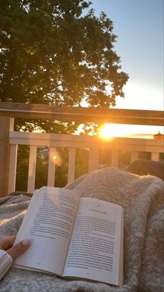 an open book sitting on top of a bed next to a wooden railing with trees in the background