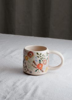 a coffee cup with flowers painted on it sitting on a white tablecloth covered surface