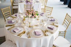 the table is set with white linens, gold chargers and purple flowers in vases