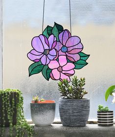 three potted plants sit on a windowsill next to a window with a stained glass flower hanging from it