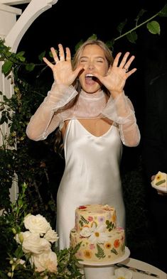 a woman in a white dress is holding her hands up to the camera while standing next to a cake