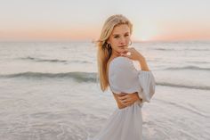 a beautiful blonde woman standing on top of a beach next to the ocean at sunset