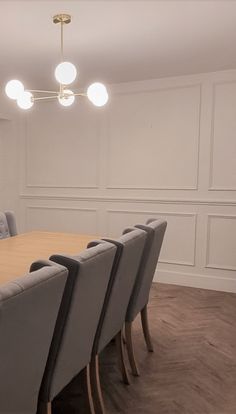 a dining room table with grey chairs and white paneled walls, along with lights above it