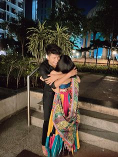 two people hugging each other in front of some trees and buildings at night with their arms around each other