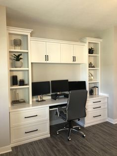 a desk with two computer monitors on top of it and white cabinets above the desk