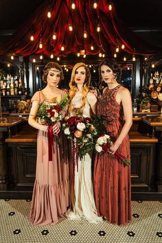 three beautiful women standing next to each other in front of a bar holding bouquets