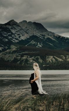 a bride and groom standing on the shore of a lake with mountains in the background