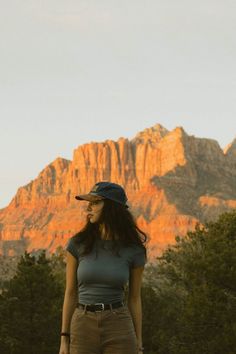 a woman standing in front of a mountain with trees and grass on the ground, wearing a hat