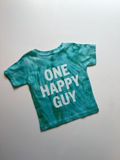 Celebrate your little one's big milestone with a touch of retro charm with our "One Happy Guy" first birthday shirt for baby boys. This tie-dye shirt is the perfect blend of fun and style for your little man's special day. Featuring a vibrant color palette and a playful retro theme, this 1st birthday boy shirt is sure to stand out in all the birthday photos and create lasting memories. Made for comfort and cuteness, this one-year-old boy shirt is a must-have birthday outfit for your little guy a Fun Short Sleeve Tops For First Birthday, Blue Cotton T-shirt For First Birthday, Fun Blue Top For First Birthday, Playful Short Sleeve Top For First Birthday, Green Graphic Print T-shirt For First Birthday, Green Cotton T-shirt For First Birthday, Cute T-shirt For First Birthday And Father's Day, Cute T-shirt For First Birthday On Father's Day, Cotton Fun Shirt For First Birthday