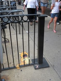 a yellow and black object on the ground behind a metal fence with people walking by