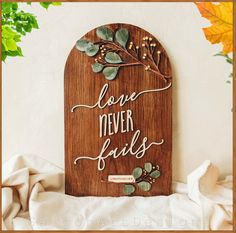 a wooden plaque with the words love never falls on it, surrounded by leaves and flowers