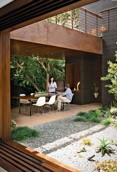 two people are sitting at a table in the backyard with plants and rocks on the ground