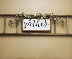 a sign that says gather hanging on the side of a wall with flowers and greenery