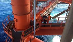 two men standing on the deck of an orange ship in the ocean with blue water