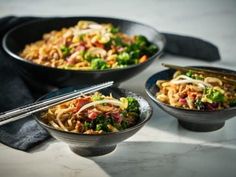 three bowls filled with noodles and vegetables on top of a white tablecloth next to chopsticks