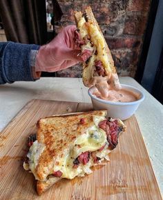 a person holding a sandwich over a wooden cutting board with a bowl of sauce in the background