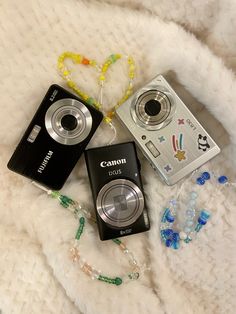 two cameras sitting on top of a white blanket next to some beads and necklaces