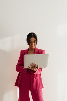 a woman in a pink suit is holding a laptop and looking at the camera while standing against a white wall