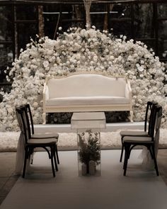 a white wedding set up with chairs and a floral arrangement on the back wall in front of it