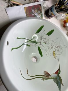 a bathroom sink with flowers painted on the bottom and green leaves in the middle, surrounded by paintbrushes