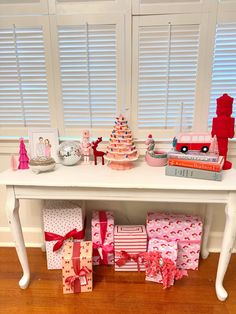 a white table topped with lots of presents under a window next to a christmas tree