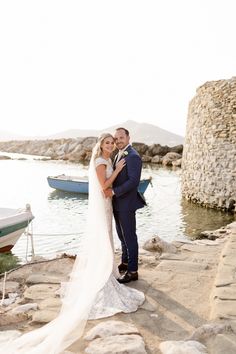 a bride and groom pose for a photo on the rocks at their wedding in front of boats