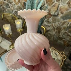 a hand holding a pink vase in front of a table with plates and candles on it