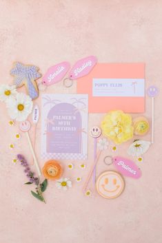 the contents of a birthday party laid out on a pink surface with flowers and paper