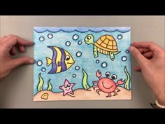 two hands holding up a drawing of an ocean scene with fish and sea animals on it