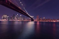 the city skyline is lit up at night as seen from across the water with a bridge in the foreground