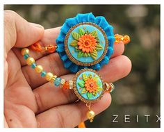 a hand holding a blue and orange flower brooch with beads on it's side