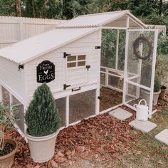 a white chicken coop in the middle of a yard with potted plants and trees