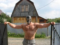 a shirtless man wearing a gas mask standing by a fence with his arms stretched out