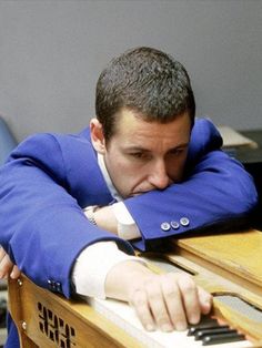 a man in a blue suit leaning his head on a wooden desk with one hand over his face