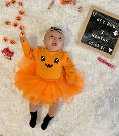 a baby dressed up as a pumpkin laying on a white rug next to halloween decorations