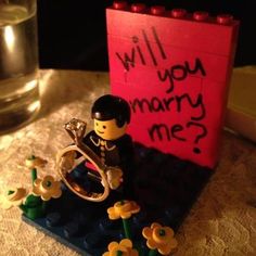 a lego figurine is sitting on a table next to a sign that says will you marry me?