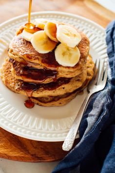 a stack of pancakes topped with bananas and syrup on a white plate next to a fork