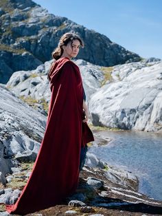 a woman in a red cape standing on rocks near water