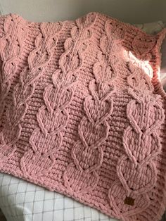 a pink knitted blanket sitting on top of a white bed next to a pillow