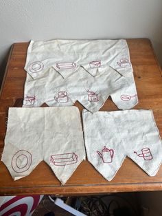 four tea towels on a wooden table with red thread embroidered onto them, and coffee mugs in the background