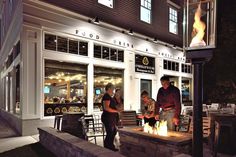 people are standing around a fire pit in front of a restaurant at night with the lights on