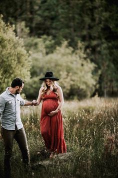 a pregnant woman in a red dress and cowboy hat holds the hand of her husband