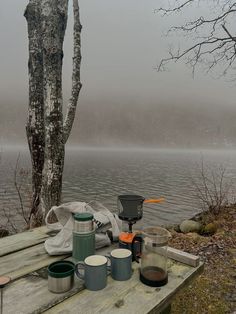 a picnic table with cups and mugs on it near the water in the fog