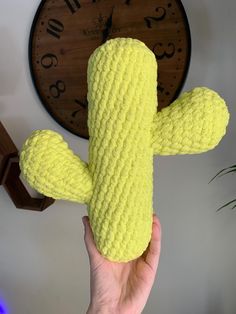 a hand holding a yellow knitted cactus toy in front of a wall mounted clock