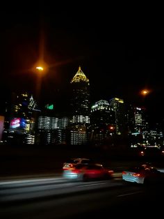 some cars are driving down the road at night in front of tall buildings and skyscrapers