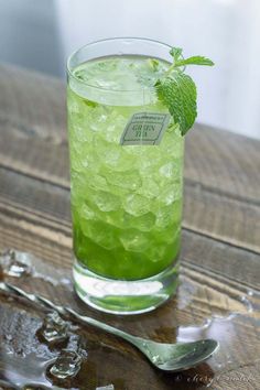 a green drink with ice and mint on a wooden table next to a silver spoon